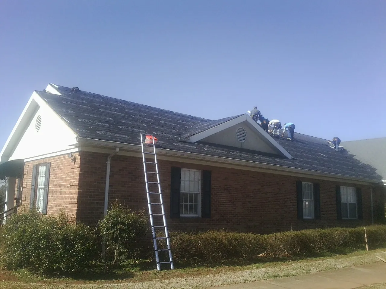 Crew tearing shingles off a brick ranch roof with a ladder set — Emergency Roof Repair work by our crew in Shallotte