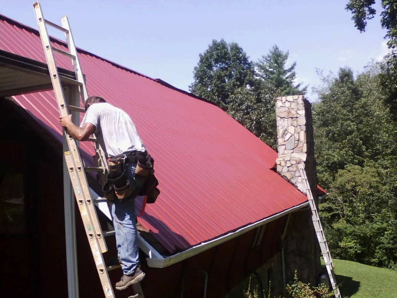 Roofer on a ladder beside a red metal roof and stone chimney — Soffit & Fascia Repair by Roofing Brothers in Shallotte